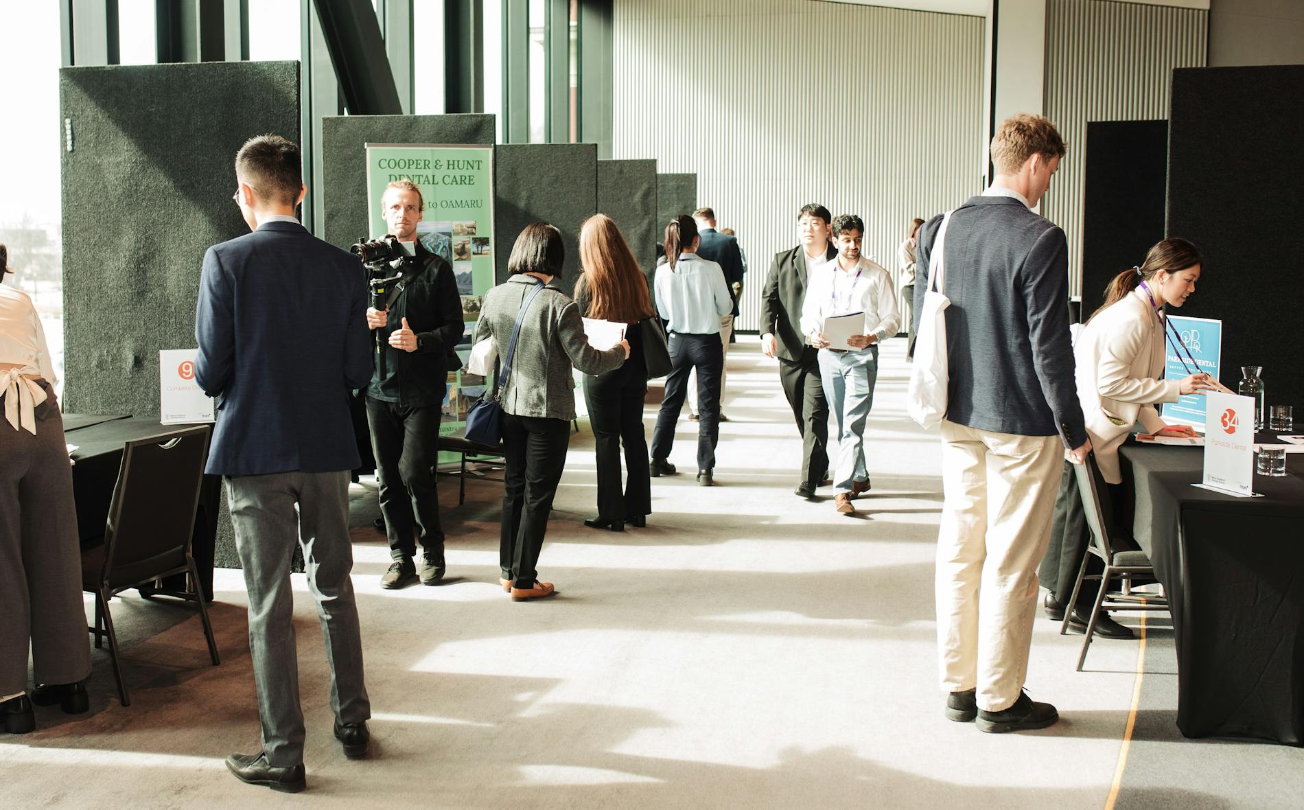 In a brightly lit venue, professionals engage in discussions at various booths, including a 'Te Pae' display, while others hold documents and converse, creating a dynamic networking environment.
